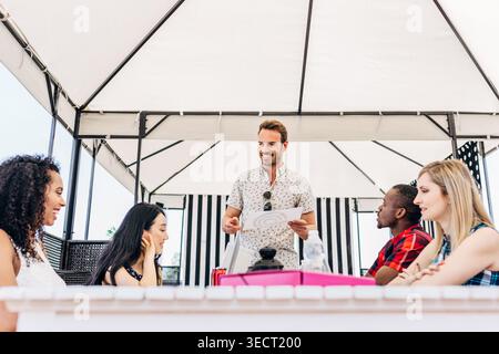 Diverse business team gathered at an outdoor table under a canopy, listening to a leader presenting documents, discussing ideas and planning a project Stock Photo