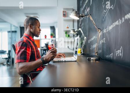 Side view of a focused freelancer in a modern workspace, typing on a laptop and holding a red takeaway cup beside a chalkboard wall, with desk lamps a Stock Photo