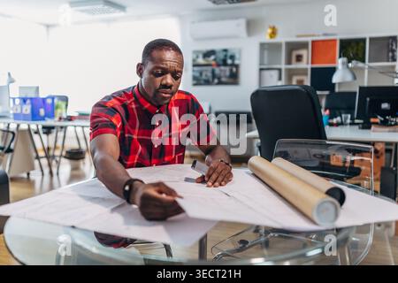 Focused male designer drafts architectural plans on a large sheet at a glass table in a bright modern office, with rolled blueprints, laptop, and supp Stock Photo