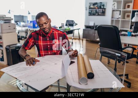 Focused designer in a red plaid shirt examines blueprint drawings at a glass desk in a bright studio office, with rolled plans and workspace equipment Stock Photo
