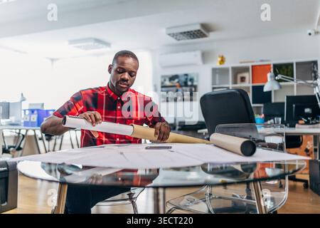 Focused young architect unrolls a large blueprint on a glass table in a modern design studio, planning a construction project with technical drawings Stock Photo