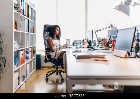 Confident professional woman sits at a desk in a bright modern office, reading on a tablet near computer monitors, paperwork, and shelves, focused on Stock Photo