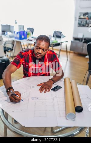 Focused young engineer draws on large architectural blueprints at a bright modern office desk, with rolled plans and smartphone nearby, planning a con Stock Photo
