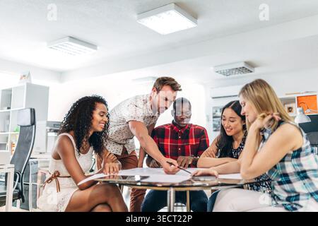 Diverse coworkers collaborate around a table in a modern office, reviewing documents and planning a project together, showing teamwork, creativity, an Stock Photo