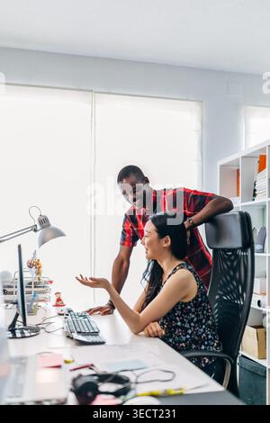 Two diverse coworkers collaborate at a bright modern office desk, discussing ideas over a computer. Casual teamwork, friendly mentoring, and productiv Stock Photo