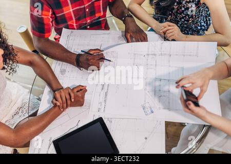 Diverse colleagues collaborate around a table, reviewing detailed architectural blueprints with pens and a tablet, planning a building project during Stock Photo