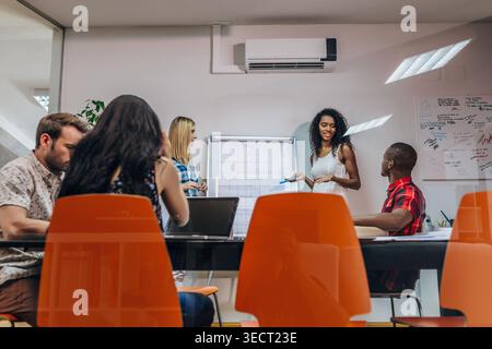 Diverse colleagues collaborate in a modern meeting room as a smiling woman presents ideas beside a whiteboard, while teammates listen, take notes, and Stock Photo