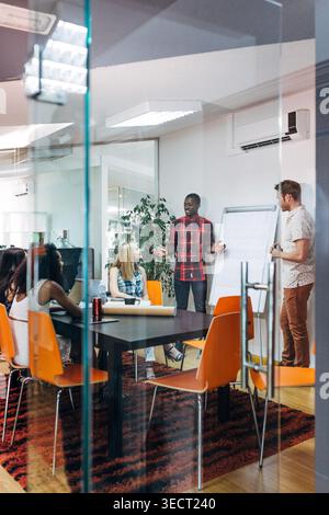 Diverse coworkers collaborate in a glass-walled conference room as a presenter leads a brainstorming meeting by a flip chart, with orange chairs and a Stock Photo