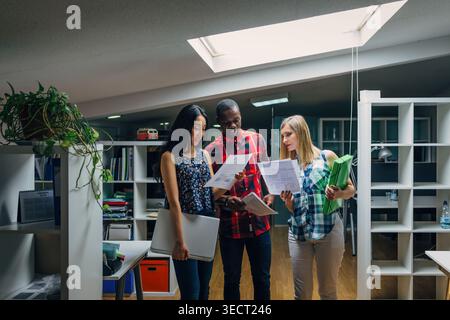 Three diverse coworkers stand in a bright modern office, discussing paperwork and sharing ideas during a collaborative meeting, with shelves, plants, Stock Photo