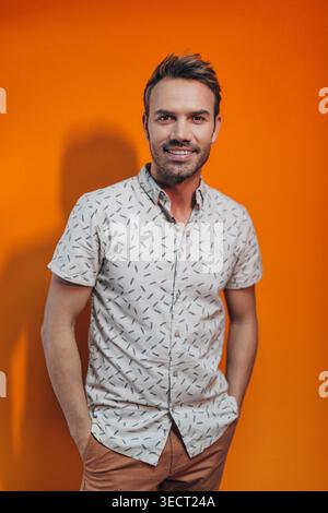 Smiling young man in patterned short sleeve shirt and brown pants poses with hands in pockets against a bright orange studio background, conveying con Stock Photo
