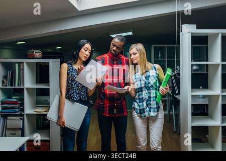 Three diverse colleagues stand in a modern open office, reviewing printed documents and folders, discussing tasks and planning a project with focused Stock Photo