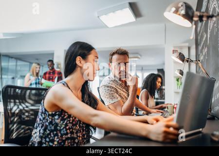 Diverse startup team collaborates in a modern open-plan office, working on laptops and discussing ideas under bright lighting, showcasing productivity Stock Photo