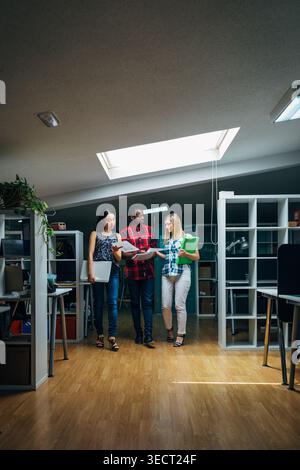 Three coworkers stand in a modern open office under skylight, reviewing printed reports and holding folders, collaborating on a project discussion wit Stock Photo