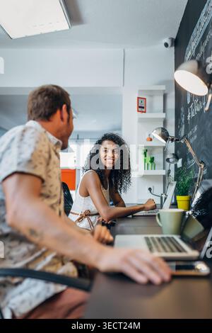 Smiling colleagues collaborate at a shared desk in a modern office, using laptops under desk lamps, with a chalkboard wall and plants creating a creat Stock Photo