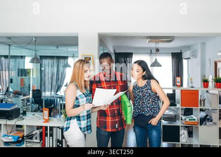 Three diverse coworkers stand together in a modern office, reviewing printed documents and a green folder, discussing plans and collaborating on a pro Stock Photo