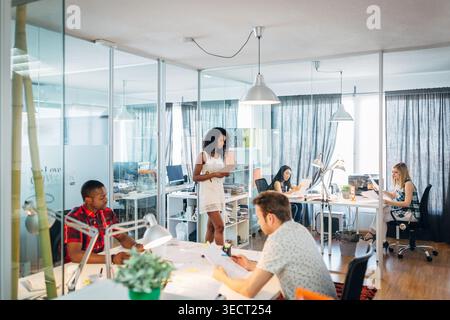 Diverse startup team collaborating in a modern open plan office with glass walls, laptops, and documents. Bright workspace with coworkers focused on t Stock Photo