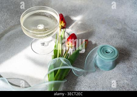 Red tulips rest on a gray tabletop beside a coupe glass and a curled spool of sheer ribbon. Bright sunlight casts soft shadows, creating a calm spring Stock Photo