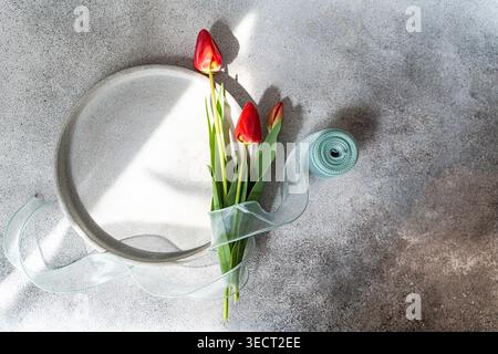 Top view of red tulips beside a simple plate and a rolled translucent ribbon on a textured gray table, creating a calm minimalist still life with soft Stock Photo