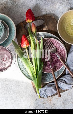 Top view flat lay of red tulips on a rustic wooden board with patterned plates, forks and knives on a light table surface, creating a cozy spring dini Stock Photo