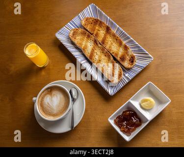 Overhead view of a morning breakfast set with toasted bread on a plate, a cappuccino with latte art, orange juice shot, and butter with fruit jam on a Stock Photo