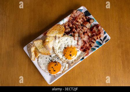 Hearty morning breakfast served on a rectangular plate with two fried eggs, crispy bacon, and sliced toast, photographed from above on a rustic wooden Stock Photo