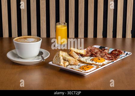 Morning breakfast on a wooden table with cappuccino, fresh orange juice, and a hearty plate of fried eggs, bacon, toast, and grilled sides, ready to e Stock Photo