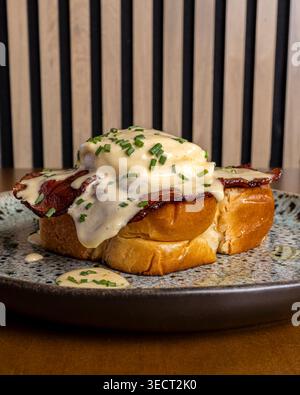 Close-up of breakfast eggs benedict served on thick toast with crispy bacon, poached egg, and creamy hollandaise sauce, topped with chives, on a rusti Stock Photo