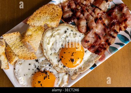 Close-up of a hearty morning breakfast plate with two sunny-side fried eggs, crispy bacon strips, and sliced rustic bread on a patterned dish, ready t Stock Photo