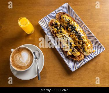 Overhead view of a morning breakfast with cappuccino in a white cup, a small glass of orange juice, and two rustic toasts topped with scrambled egg an Stock Photo