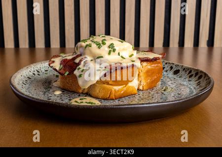 Close-up of breakfast toast topped with crispy bacon, a poached egg, and creamy hollandaise sauce, garnished with chives, served on a ceramic plate in Stock Photo