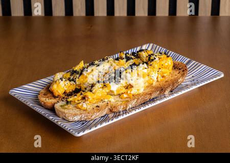 Close-up of scrambled eggs on toasted bread served on a striped rectangular plate, topped with seasoning, on a wooden table for a simple morning break Stock Photo