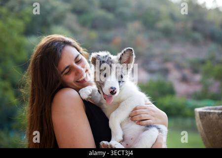 Smiling woman cuddles a playful border collie puppy in her arms outdoors, capturing a warm pet bond, soft natural light, and a relaxed countryside bac Stock Photo