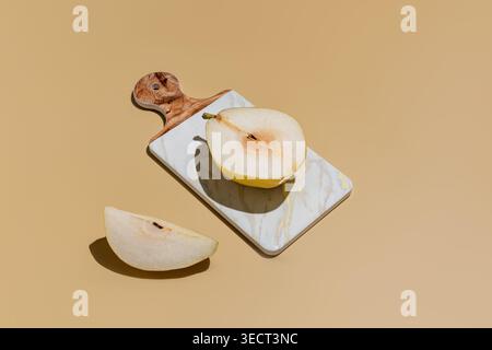 Minimal still life of ripe pear half on a marble cutting board with wooden handle, set on beige background with strong shadow. Stock Photo