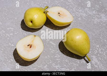 Minimal still life of fresh yellow pears, including whole fruit and sliced halves with seeds, arranged on a gray stone surface in bright natural light Stock Photo