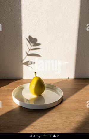 Minimal still life with a ripe yellow pear on a round white tray on wooden table, lit by warm morning sunlight and leaf shadow on textured wall, calm Stock Photo