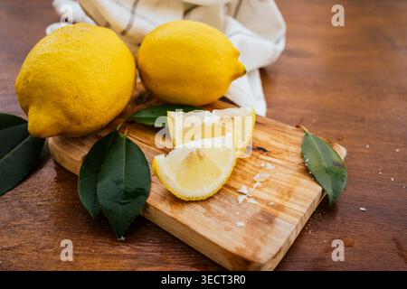 Fresh lemons with a sliced wedge on a wooden cutting board, green leaves and scattered salt on a rustic table. Bright yellow citrus fruit food for coo Stock Photo