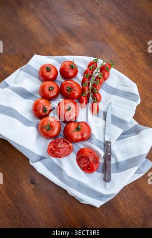 Top view of ripe red tomatoes, including cherry tomatoes on the vine, arranged on a striped kitchen towel on a rustic wooden table with a knife and sl Stock Photo