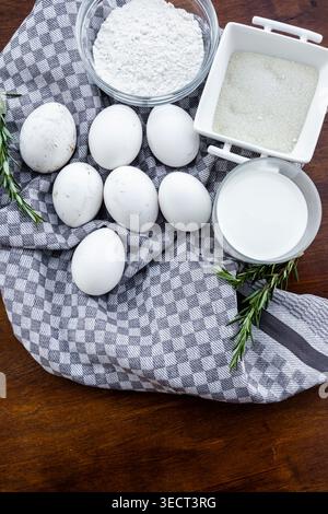 Top view of fresh eggs with bowls of flour, sugar and milk on a checkered kitchen towel with rosemary sprigs on a wooden table, rustic baking concept. Stock Photo