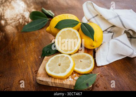 Fresh whole lemons and sliced rounds on a wooden cutting board with green leaves, coarse salt, and a kitchen towel, styled as rustic food preparation. Stock Photo