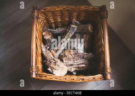 Top view of a rustic wicker basket filled with dry firewood logs on a dark wooden floor, cozy home heating concept with natural texture and warm tones Stock Photo