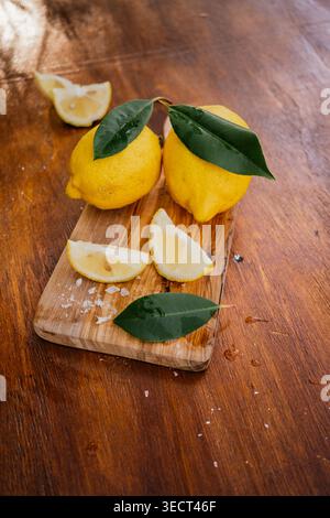 Two fresh lemons with green leaves on a wooden cutting board, with cut wedges and sea salt on a rustic table. Natural citrus food styling for cooking Stock Photo