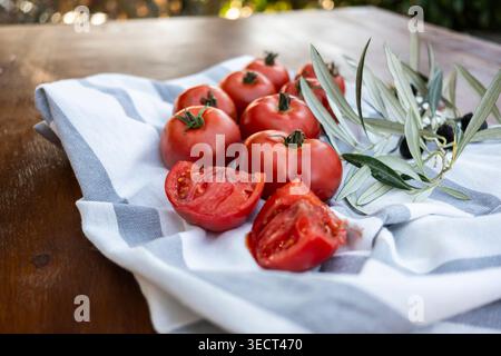 Ripe red tomatoes, some sliced open, resting on a striped linen cloth beside an olive branch on a wooden table in soft natural light, ideal for Medite Stock Photo