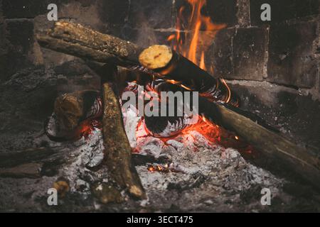Close-up of burning firewood logs in a rustic fireplace, with glowing embers, ash, and rising smoke. Warm flames create a cozy winter atmosphere and h Stock Photo