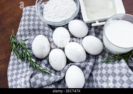 Top view of fresh white eggs with bowls of flour, sugar and milk on a checkered kitchen towel, decorated with rosemary on a wooden table for baking pr Stock Photo