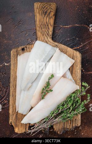 Raw cod or hake fillets with thyme on wooden board and pepper on dark textured background. Fresh pollock whitefish prepared for healthy seafood cookin Stock Photo