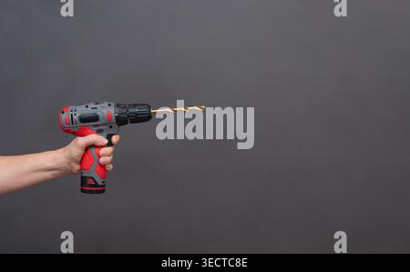 Cordless drill held in hand, red and black tool with drill bit extended, isolated on gray background. Hand holding cordless drill, power tool with met Stock Photo