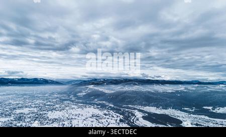 High altitude panorama of a snowy town and mountain range beneath dense layered clouds. Blue grey tones mix with rising valley fog to create a broad Stock Photo