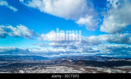 Expansive sky fills the frame with sunlit patches and towering clouds above a broad winter valley and distant ranges. Villages and fields rest Stock Photo