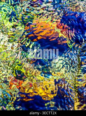 Sunlit water ripples over vivid autumn leaves in a shallow stream, blending blue, gold and amber into a luminous natural abstract background scene. Stock Photo