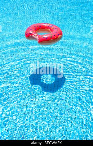 Pink donut inflatable ring floating in blue swimming pool casting soft shadow on bottom. Clean summer concept with copy space. Leisure, relaxation and Stock Photo